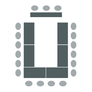 Room arranged with tables pushed together in a "U" shape with chairs around the outside edges facing a speaker panel table at the front of the room.