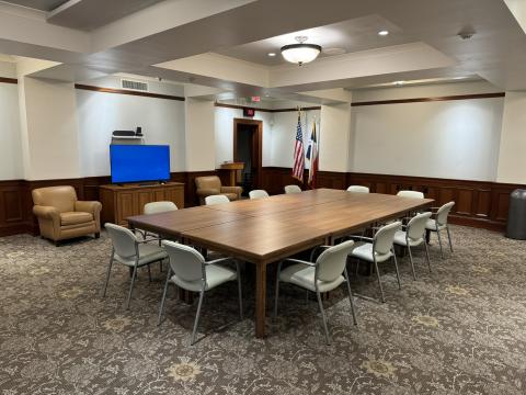 Large room with 8 square tables in the center surrounded by chairs. Large tv monitor in the background.